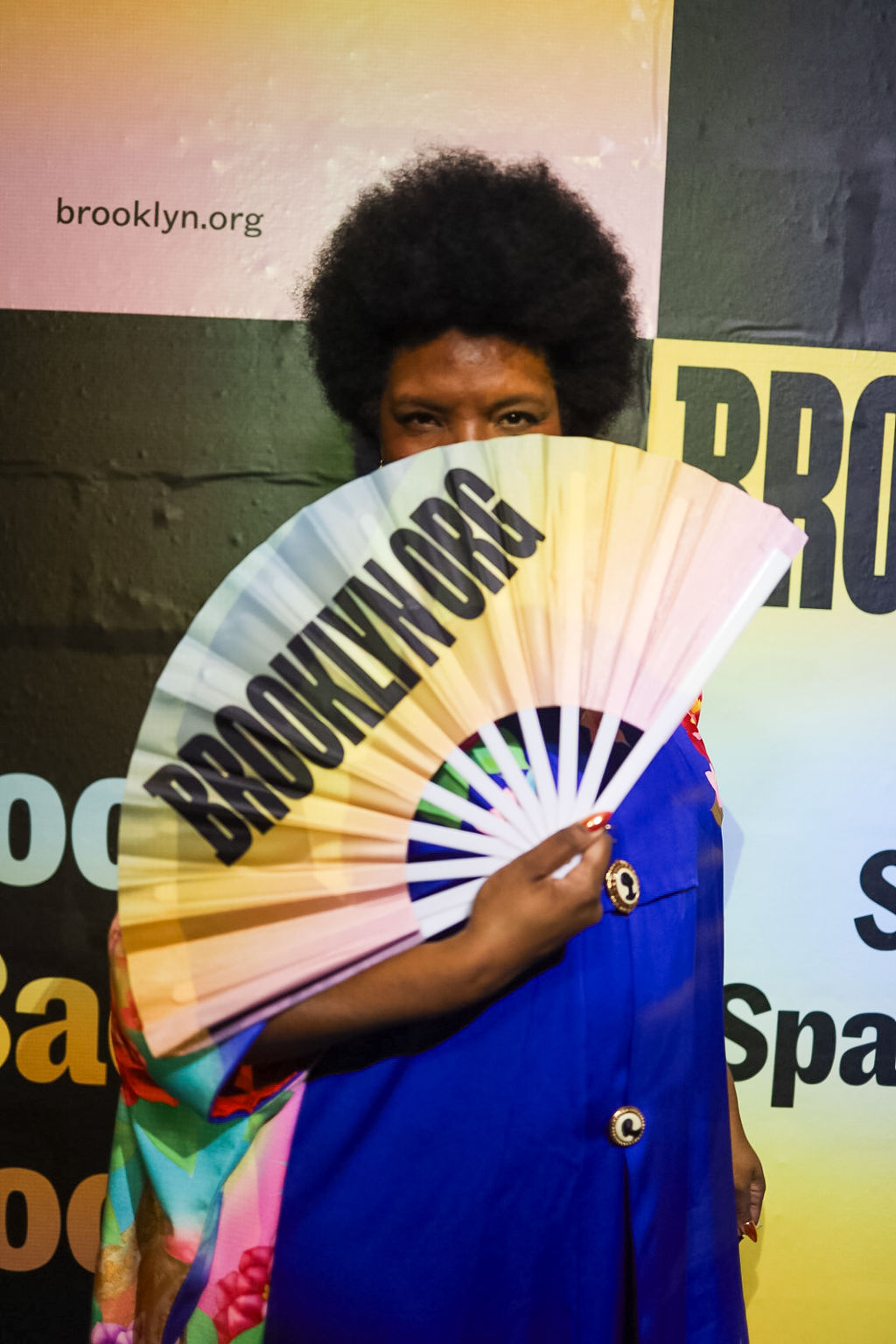 A person in a blue outfit holds a large fan with "BROOKLYN.ORG" printed on it, partially covering their face, standing in front of a yellow and black backdrop.