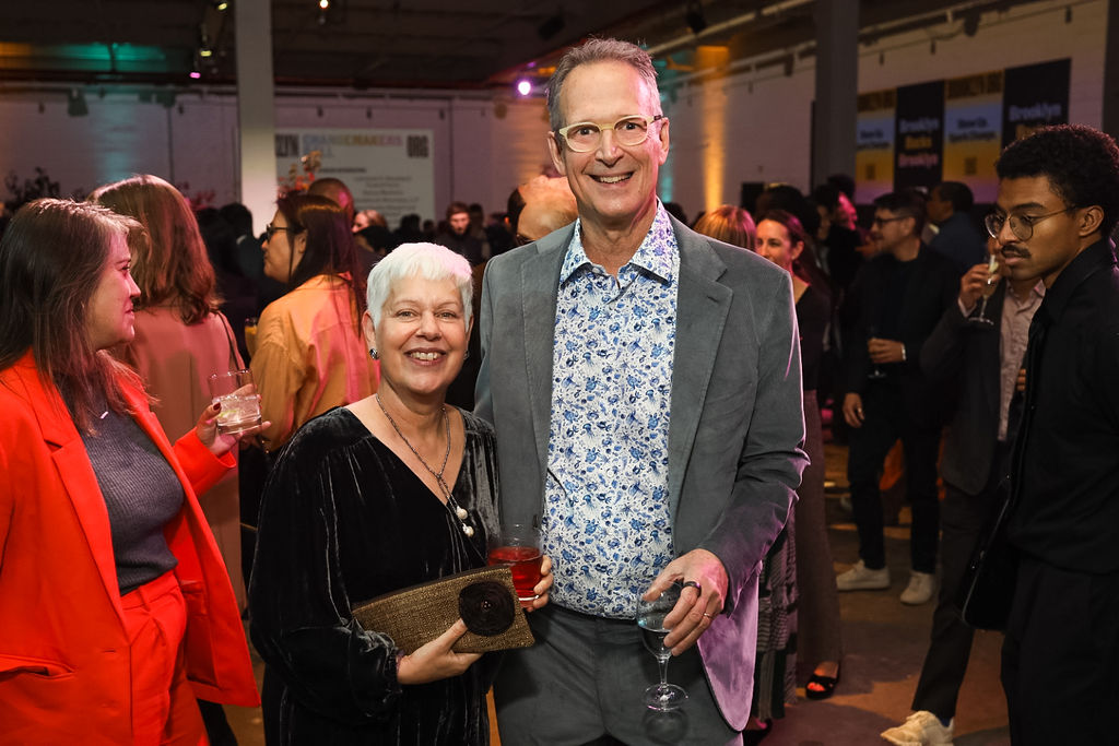 A woman and a man pose and smile for the camera at a crowded indoor social event, with people mingling in the background.