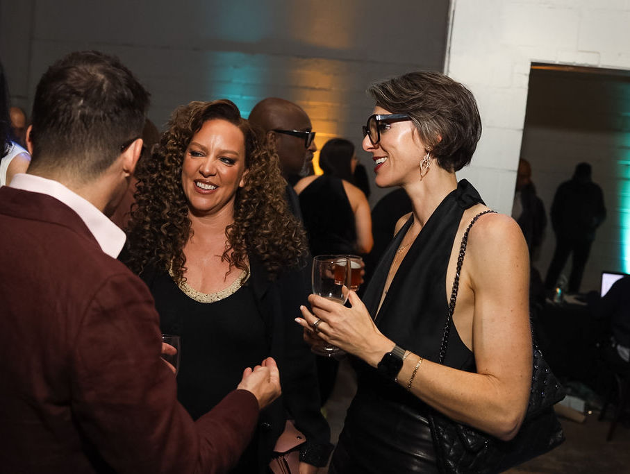 Three adults dressed in evening attire stand together at an indoor event, conversing and holding drinks.