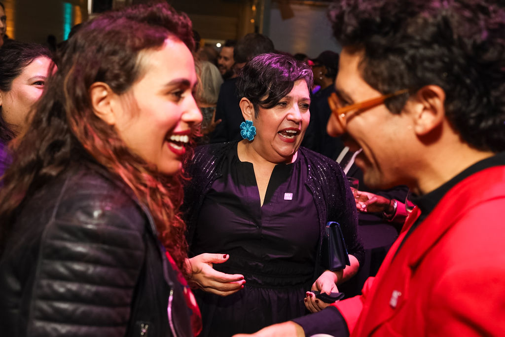 Three people stand close together at an indoor event, smiling and talking. The woman in the center wears a black dress and blue earrings, while the others wear a red blazer and a leather jacket.