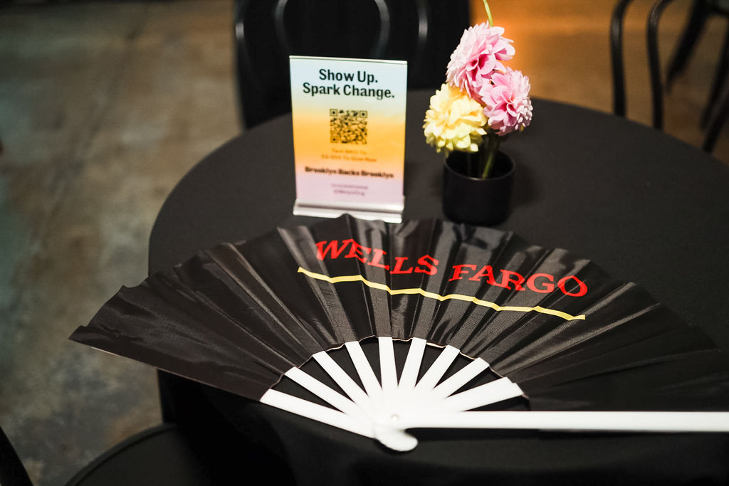 A black table holds a Wells Fargo-branded fan, a small vase with pink and yellow flowers, and a sign that reads "Show Up. Spark Change.