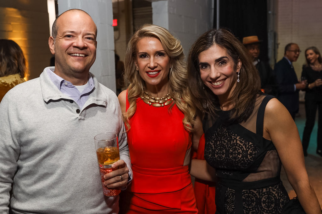 Three adults pose and smile at an indoor event; the man holds a drink, the women wear a red dress and a black dress. Other people are visible in the background.
