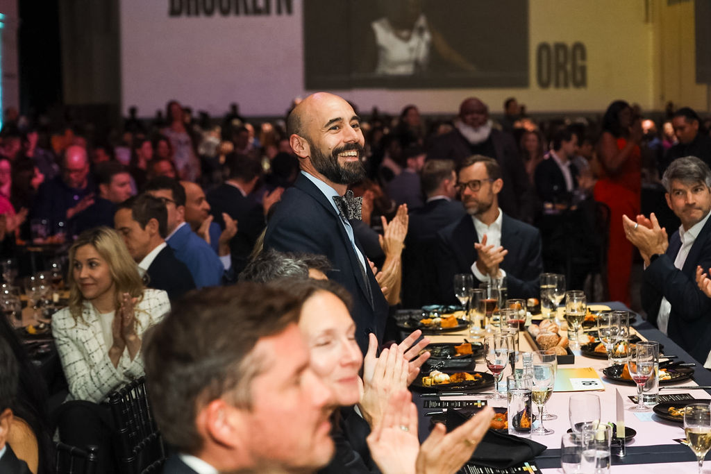 A man in formal attire stands and smiles while people seated around him applaud at a banquet event. Tables are set with food and drinks, and the room is filled with attendees.