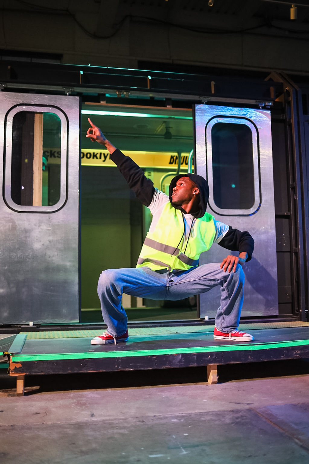 A person in a safety vest and jeans strikes a dynamic pose in front of an open subway train door on a platform.