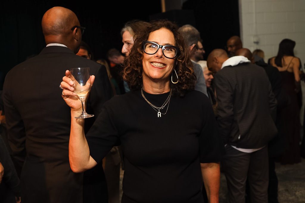 Woman with curly hair and glasses smiles while holding a wine glass at a crowded indoor event. People are gathered and socializing in the background.
