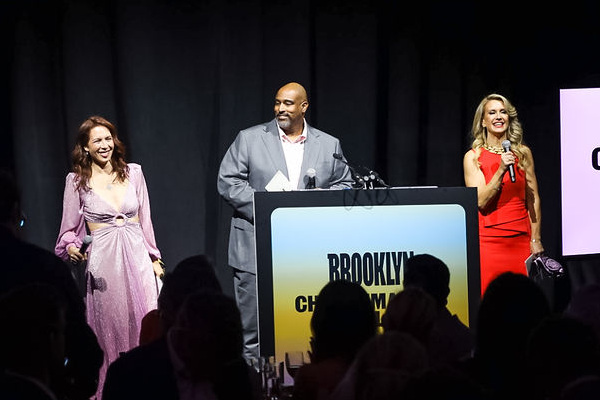 Three presenters stand on stage at the Changemakers Ball, with two large screens displaying the event name in the background.