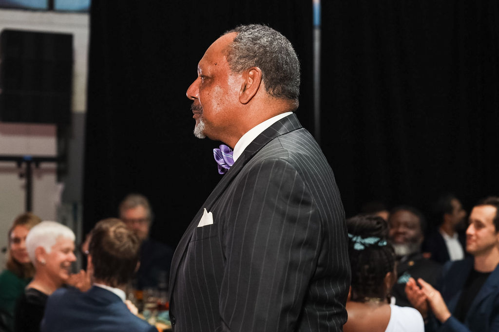 A man in a pinstripe suit and bow tie stands in profile at a formal event, with seated attendees visible in the background.