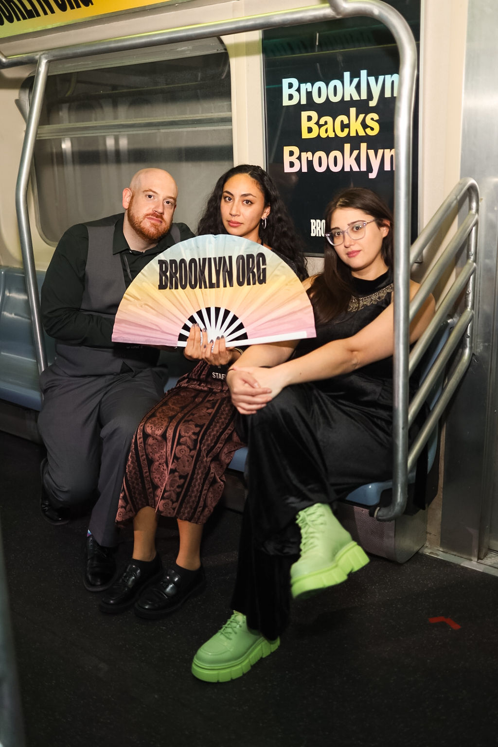Three people sit on a subway bench; one holds a fan reading "BROOKLYN.ORG." Behind them is a sign that says "Brooklyn Backs Brooklyn.