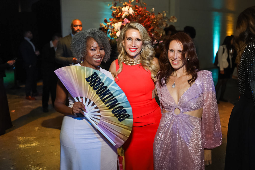 Three women pose together at an event; one holds a large fan that reads "BROOKLYN.ORG." There are people and a large floral arrangement in the background.