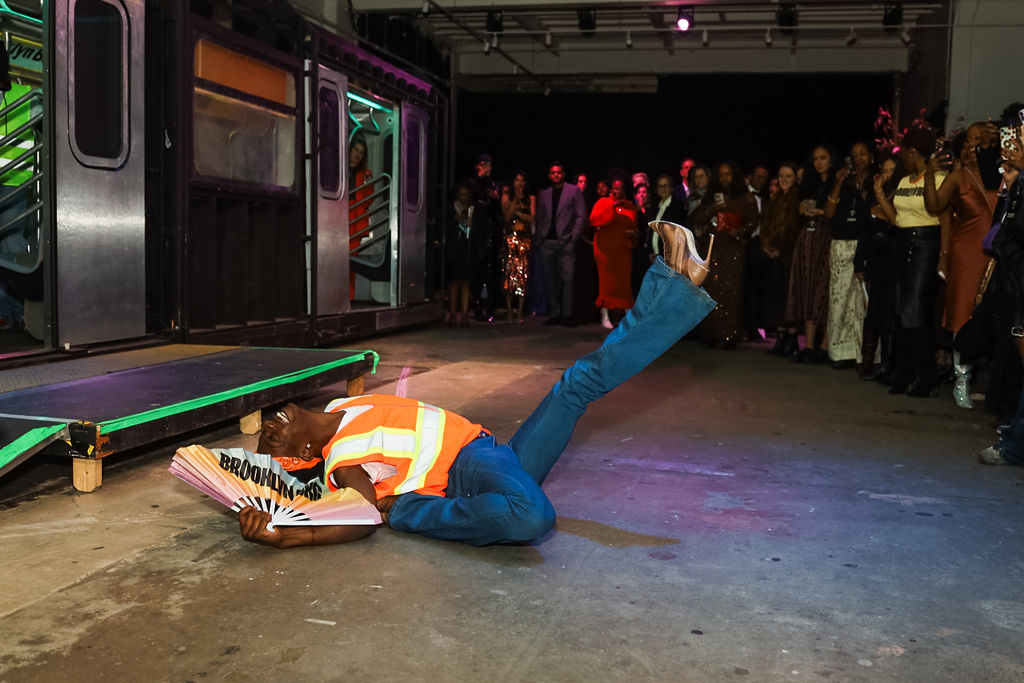 A performer in a safety vest and heels poses on the floor with one leg raised, holding a fan that reads "BROOKLYN," while a crowd watches near a subway train set.