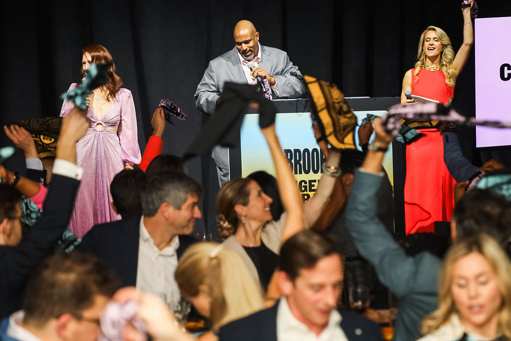 A man speaks at a podium on stage while two women stand beside him and people in the audience raise scarves in the air.