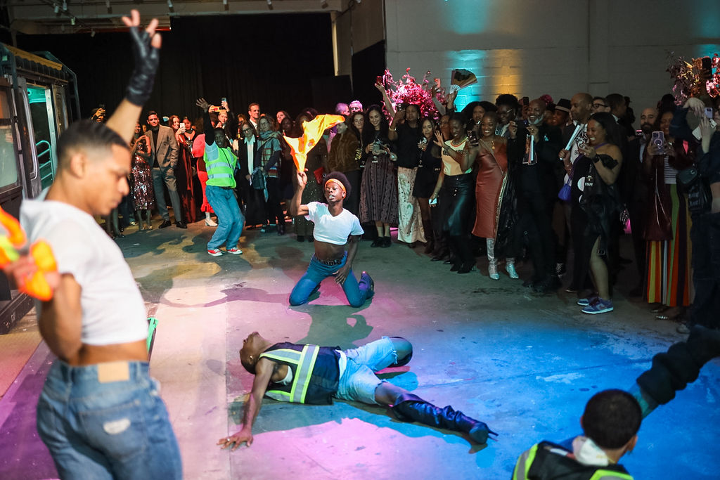A group of performers dance energetically on the floor in front of a cheering audience in a large indoor space, with colorful lighting.