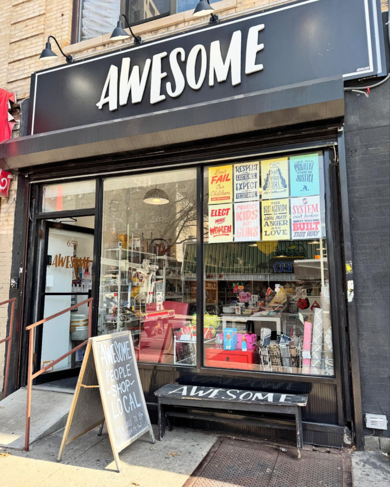 Street view of a shop named “AWESOME” with a sandwich board reading "AWESOME PEOPLE SHOP LOCAL" and various colorful products and posters visible through the window.