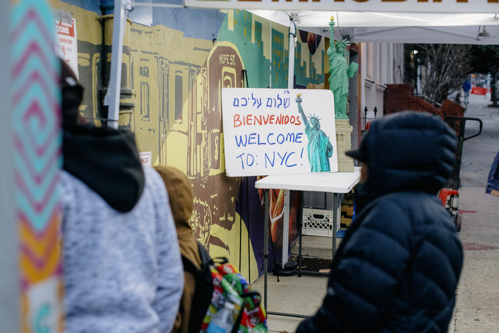 A hand-drawn sign reading "Welcome to NYC!" in English, Spanish, and Hebrew is displayed near a sidewalk, with several people in jackets walking nearby.