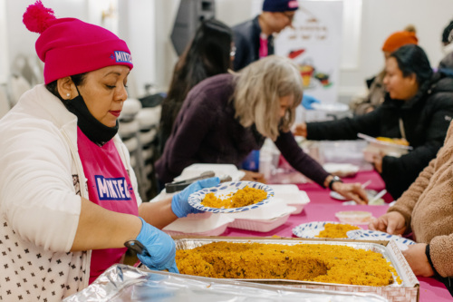 A woman in a pink hat and gloves serves rice from a tray onto a paper plate at a community event, with others serving food beside her.