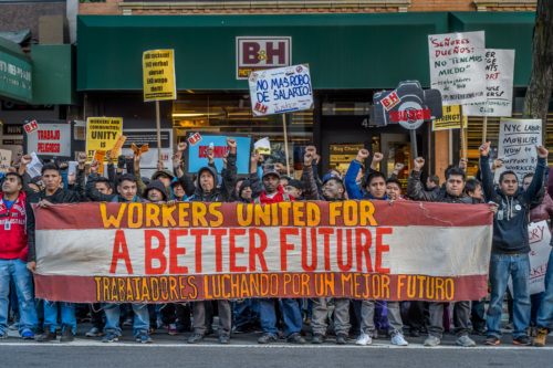 A group of workers stand in front of a store holding signs and a banner that reads, "Workers United for a Better Future" in English and Spanish.