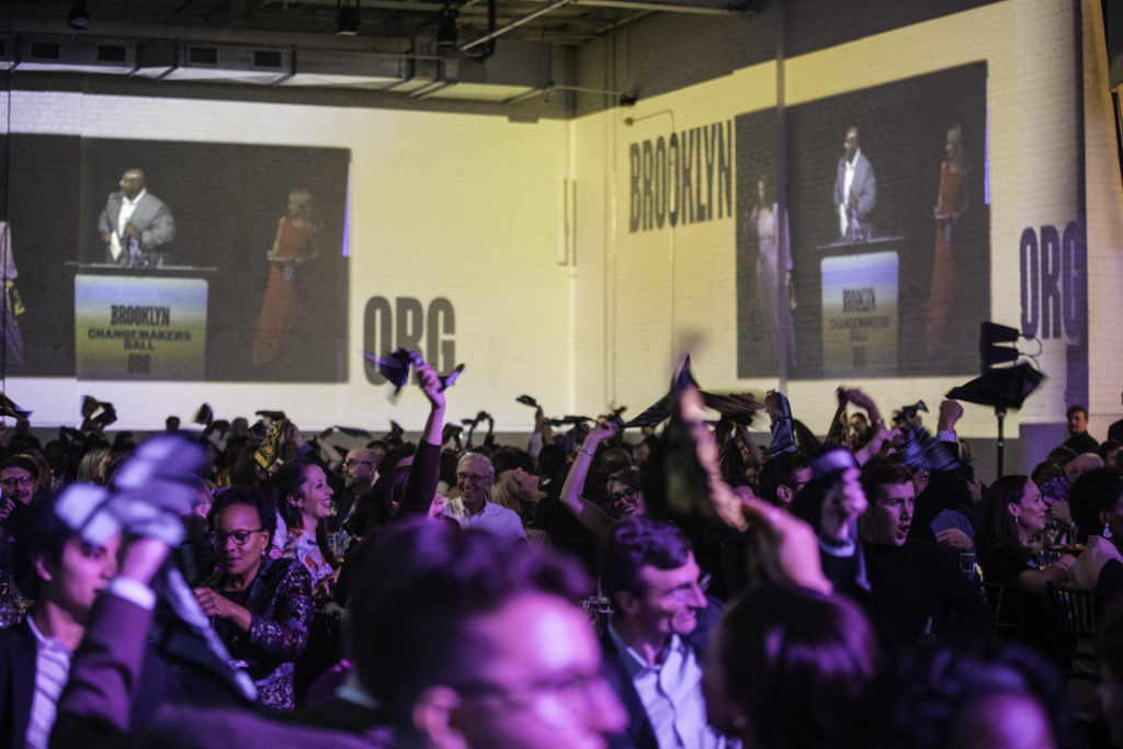 A crowd in an indoor venue waves objects in the air while a man and woman speak onstage, projected onto a large wall behind them with the word "BROOKLYN" displayed.
