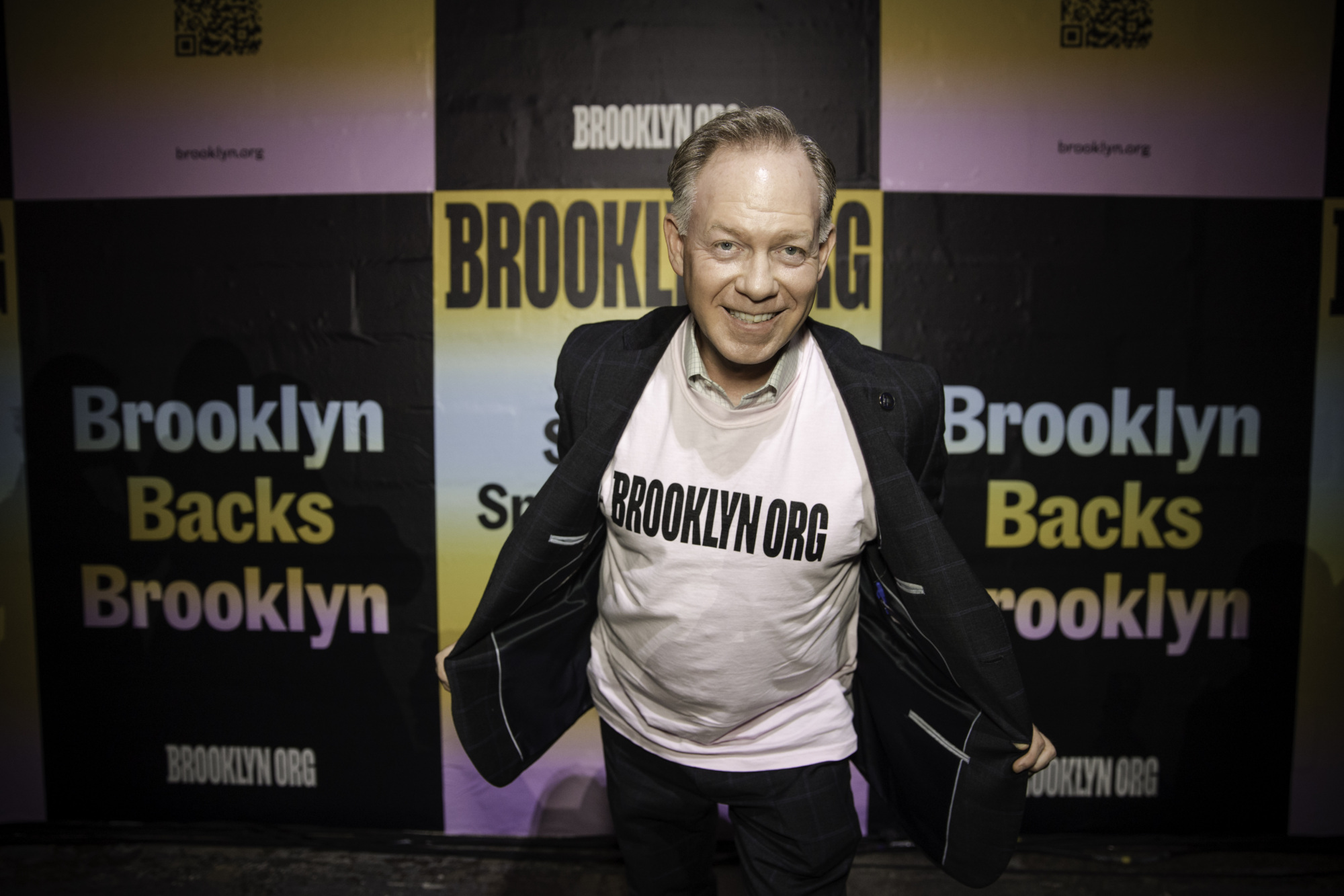 A man smiles at the camera, opening his jacket to reveal a "BROOKLYN.ORG" t-shirt, standing in front of a Brooklyn.org promotional backdrop.