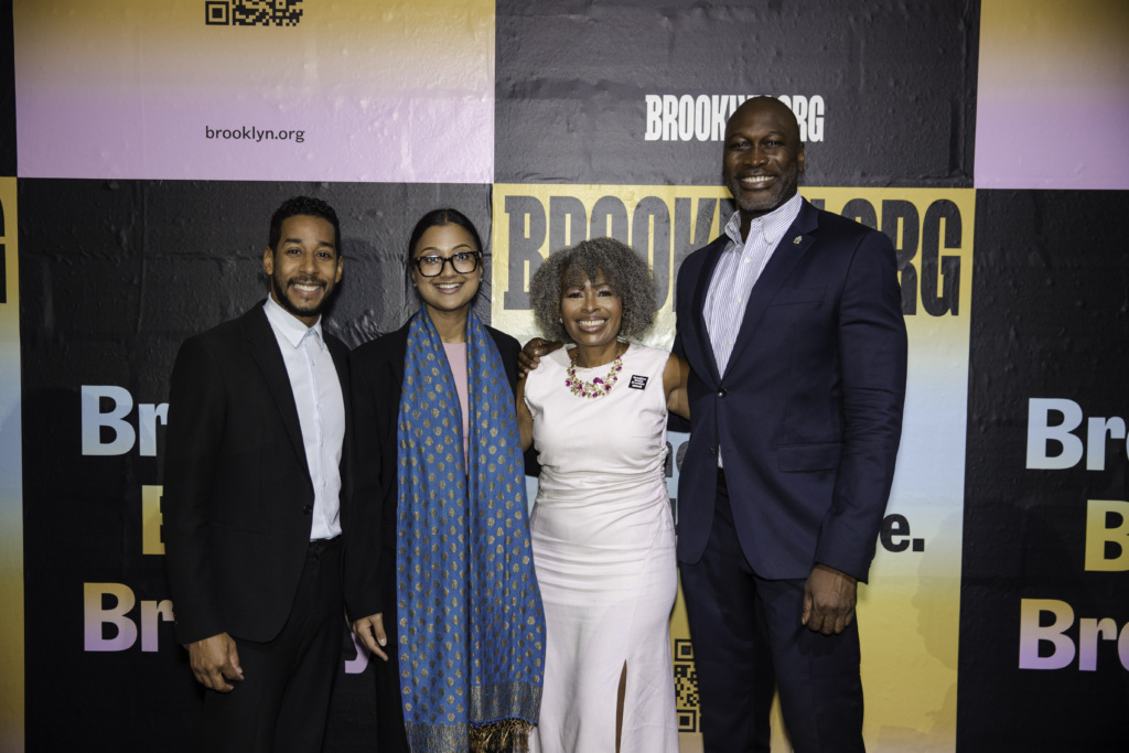 Four people stand side by side, smiling, in front of a colorful backdrop with "Brooklyn.org" and "BRIC Celebrate Brooklyn!" logos.