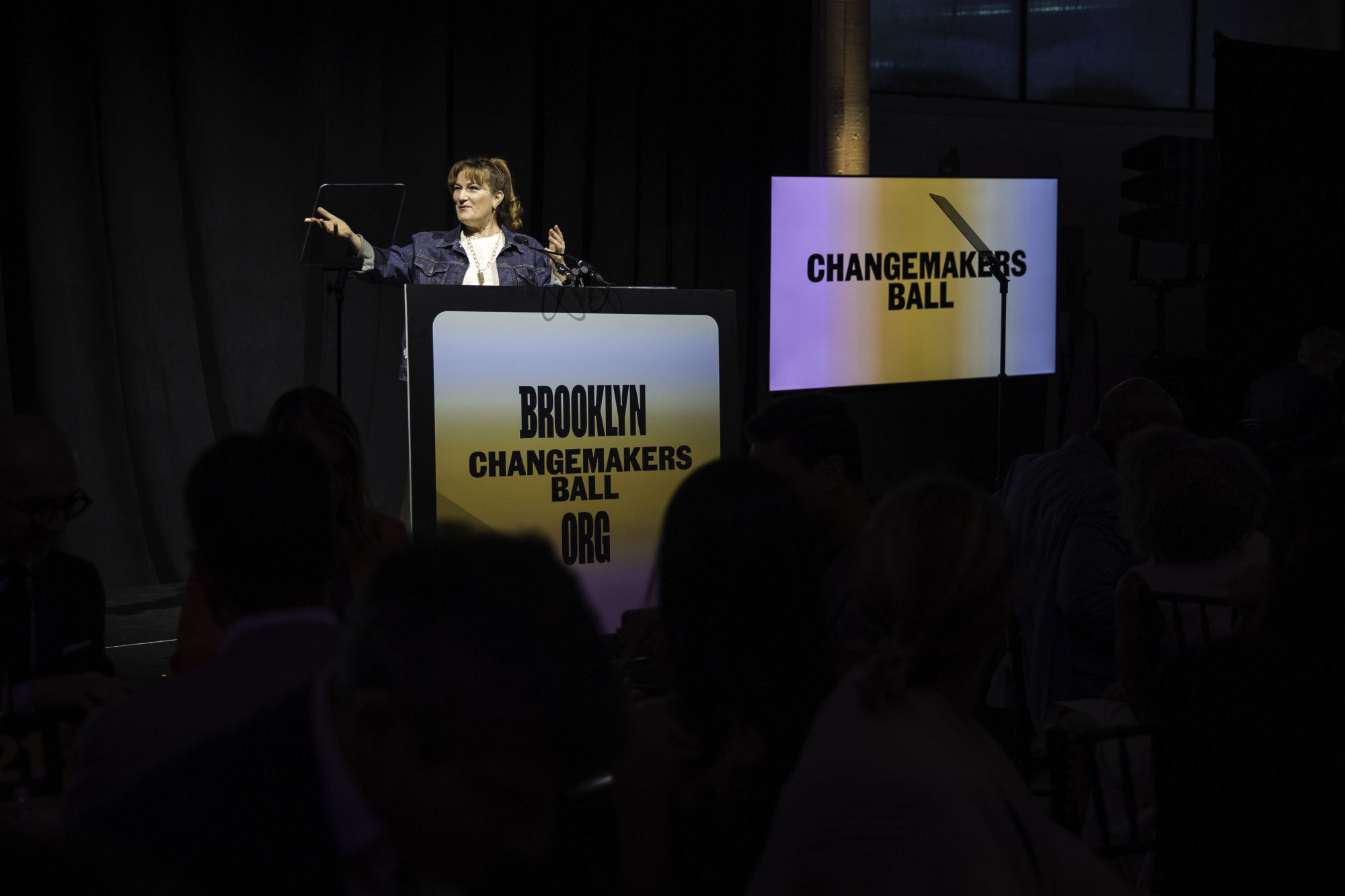 A person speaks at a podium labeled "Brooklyn Changemakers Ball.org" with a matching display screen in the background at an indoor event.
