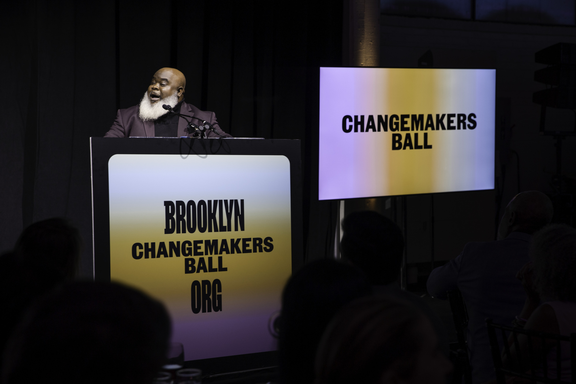 A person with a white beard speaks at a podium labeled “Brooklyn Changemakers Ball” with a matching “Changemakers Ball” sign on a screen beside them.