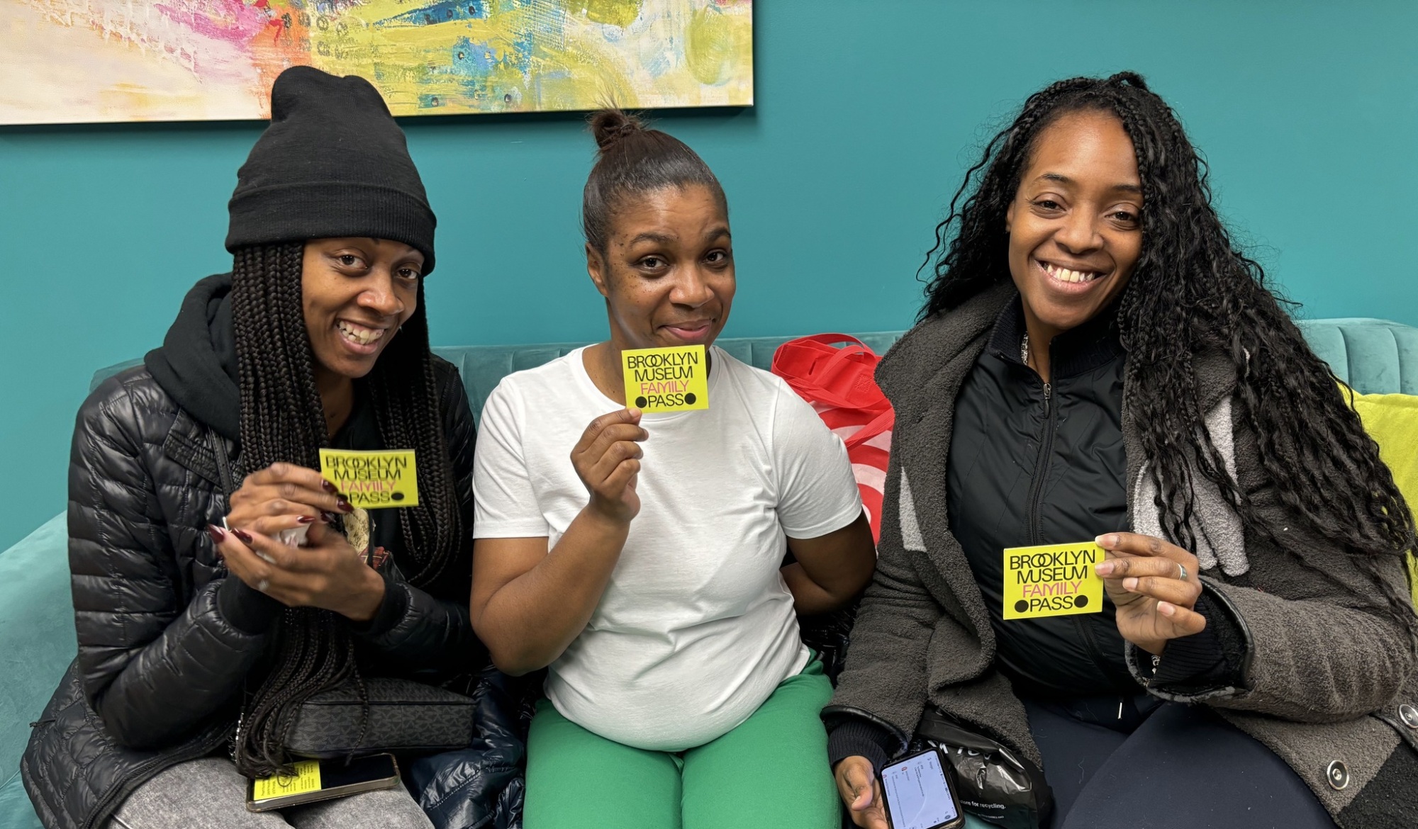 Three women sitting on a couch, smiling and holding Brooklyn Museum passes; one woman holds a mobile phone in her lap.
