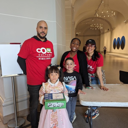 Four adults and two children pose together at an indoor event table. The adults wear matching red "COOL CULTURE" shirts, and the girl in front wears a traditional dress.