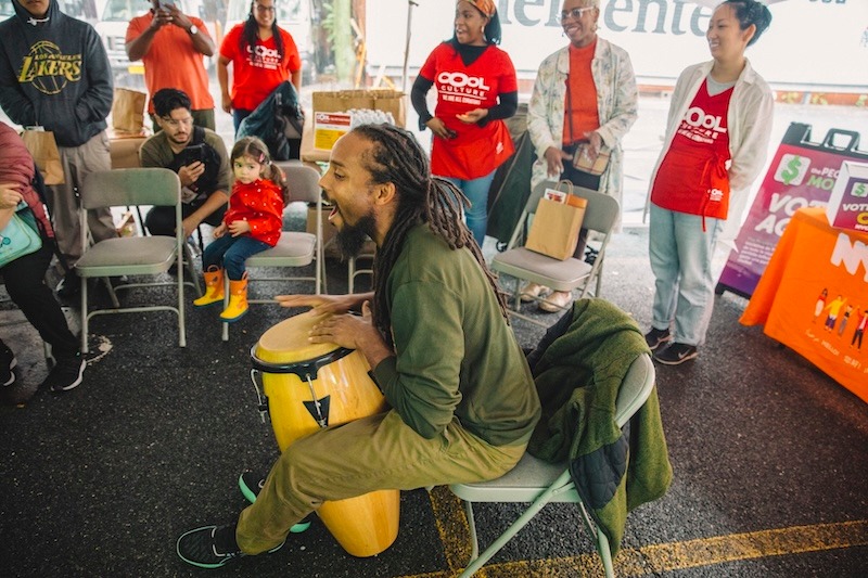 A man plays a drum while seated, surrounded by adults and children, some wearing red shirts with "Cool Culture" written on them, at an outdoor community event.