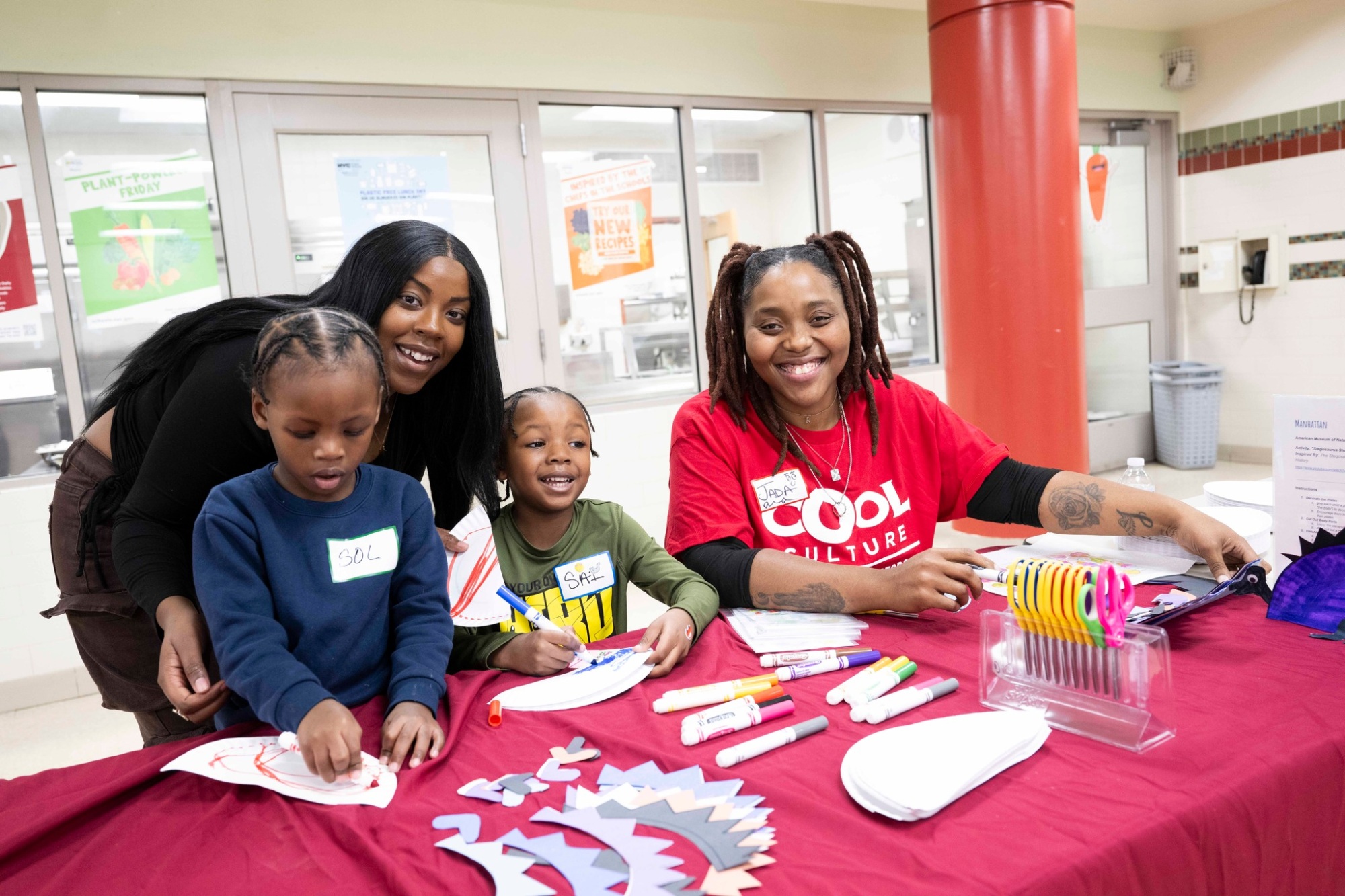 Two adults and two children sit at a table with arts and crafts supplies, smiling at the camera in a community center setting.
