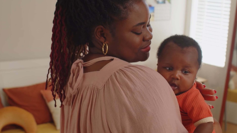 A woman holds a baby over her shoulder in a softly lit room with a bed and cushions in the background.