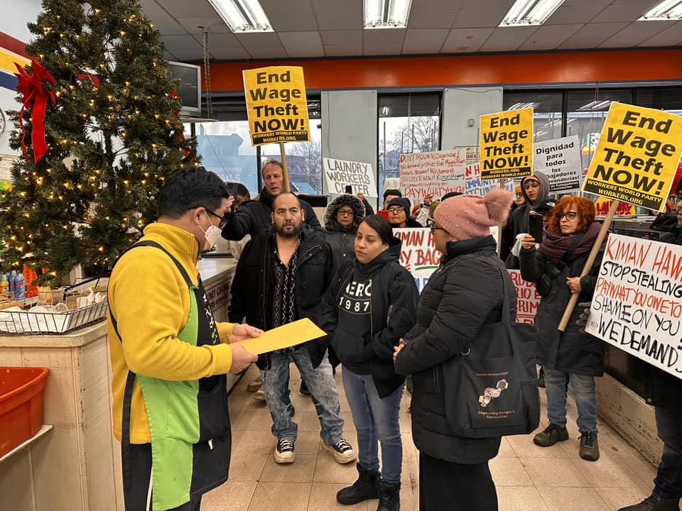 A group of people inside a laundromat hold signs reading "End Wage Theft Now!" while one person hands a yellow envelope to a laundromat worker.