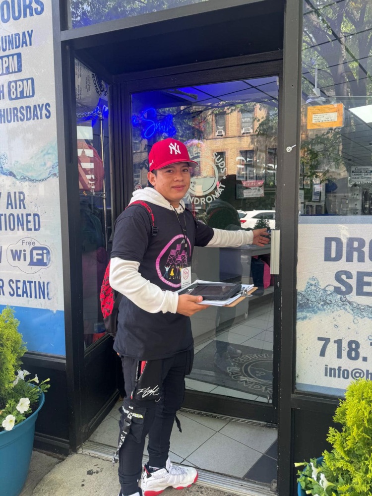 A person in a red cap holds food containers and opens the glass door of a laundromat, with signage visible on the storefront windows.