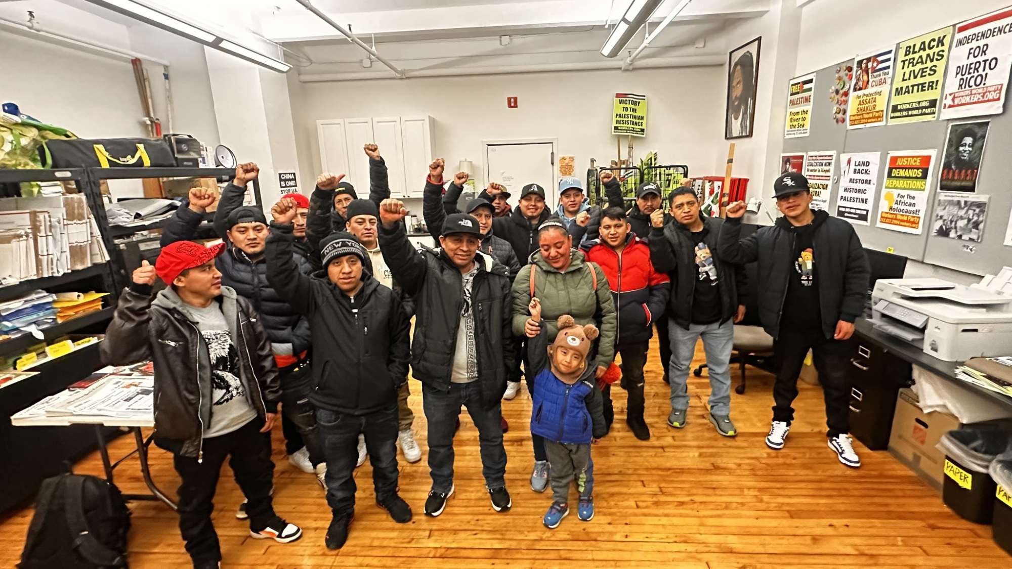 A group of people, including adults and children, stand together indoors with some raising their fists; activist posters are visible on the walls in the background.