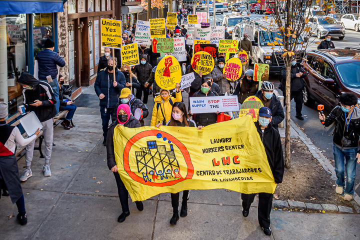A group of people march on a city sidewalk holding signs and a yellow Laundry Workers Center banner during a protest.