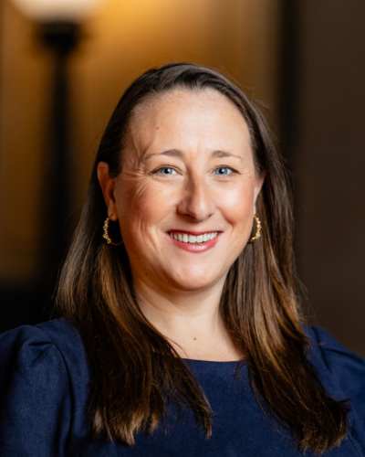 A woman with long brown hair wearing a navy blue dress and gold hoop earrings smiles at the camera against a blurred indoor background.