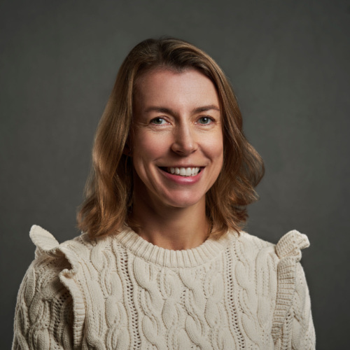 Woman with shoulder-length light brown hair wearing a cream-colored cable-knit sweater, smiling in front of a plain dark background.