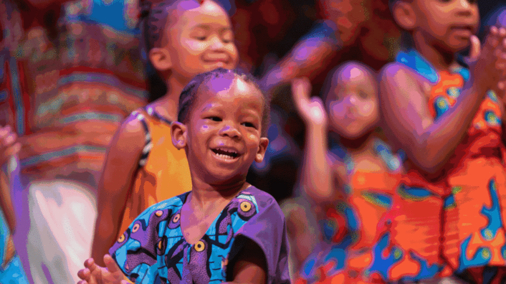 A young child smiles and claps, surrounded by other children in brightly colored clothing, during a lively group activity or performance.