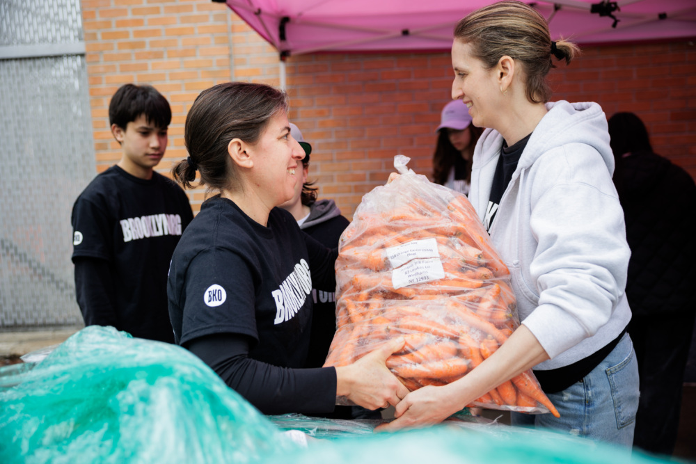 Two people exchange a large bag of carrots at an outdoor event, with others in the background and a pink canopy overhead.