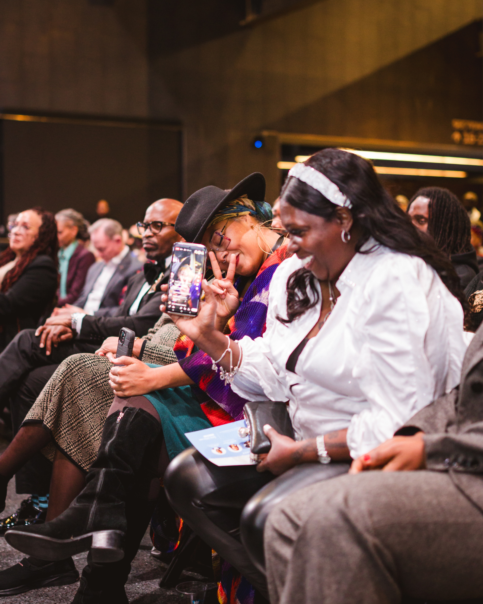 Several people sit in an audience; a woman in the foreground smiles and holds up her phone, appearing to take a photo or video.