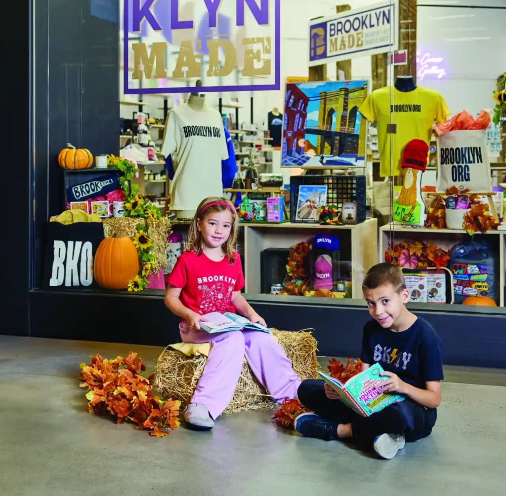 Two children sit on hay bales reading books in front of a Brooklyn Made store display decorated with pumpkins, leaves, and Brooklyn-themed merchandise.