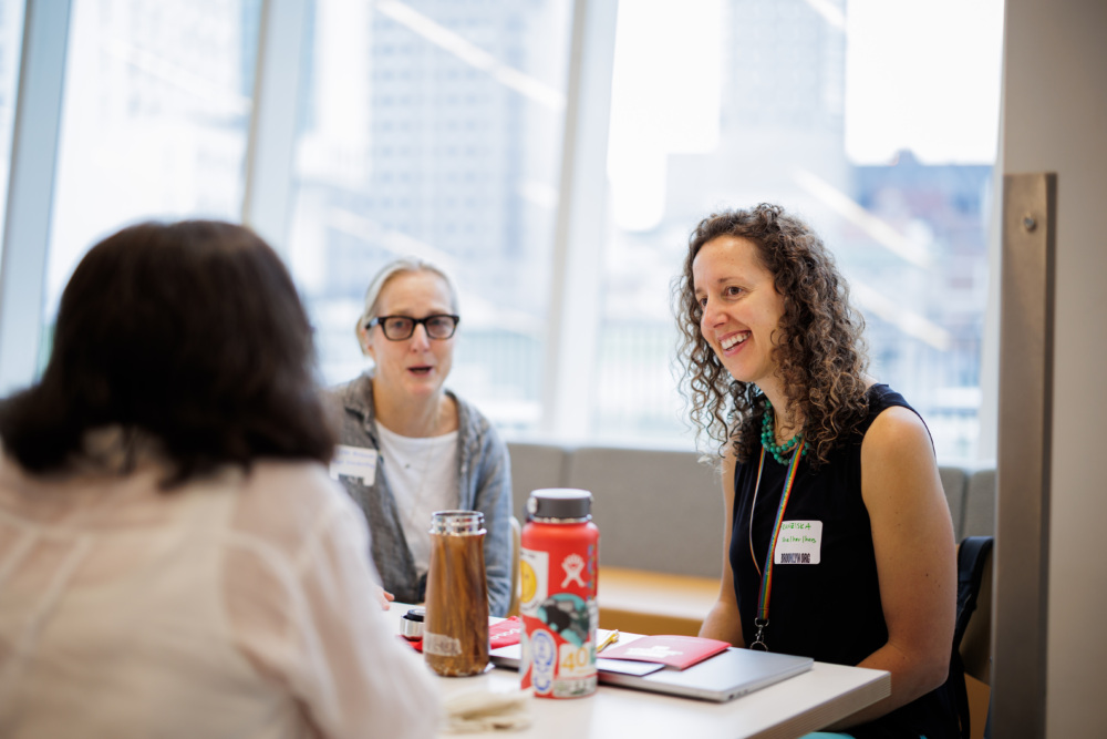 Three women sit at a table in a bright, modern office, talking. Two wear name tags, one smiles, and notebooks and water bottles are on the table.