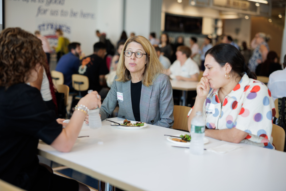 Three women sit at a table eating lunch and conversing in a busy cafeteria or event space with people in the background.