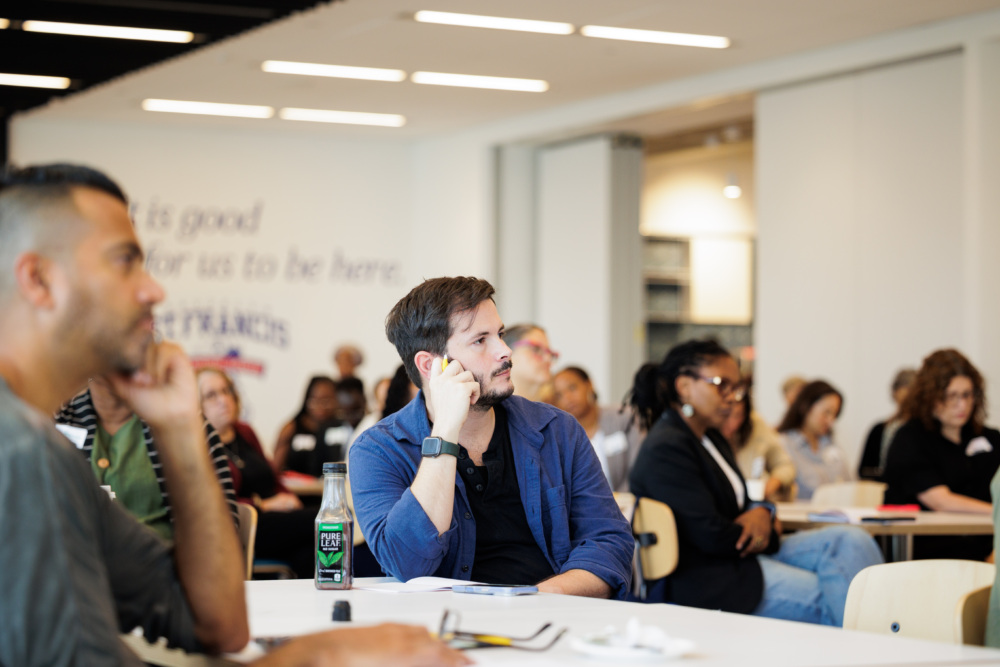 A group of people sit at tables in a modern conference room, attentively listening to a presentation. One man in the foreground rests his chin on his hand.