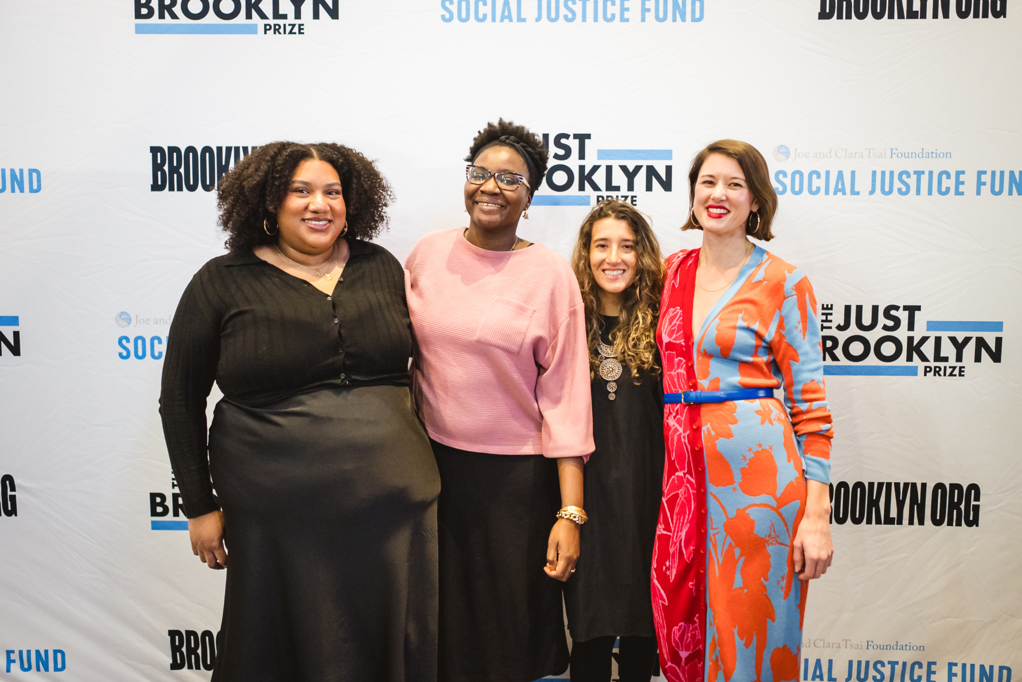 Four women stand together, smiling, in front of a step-and-repeat banner with "Brooklyn", "The Just Brooklyn Prize", and "Social Justice Fund" logos.