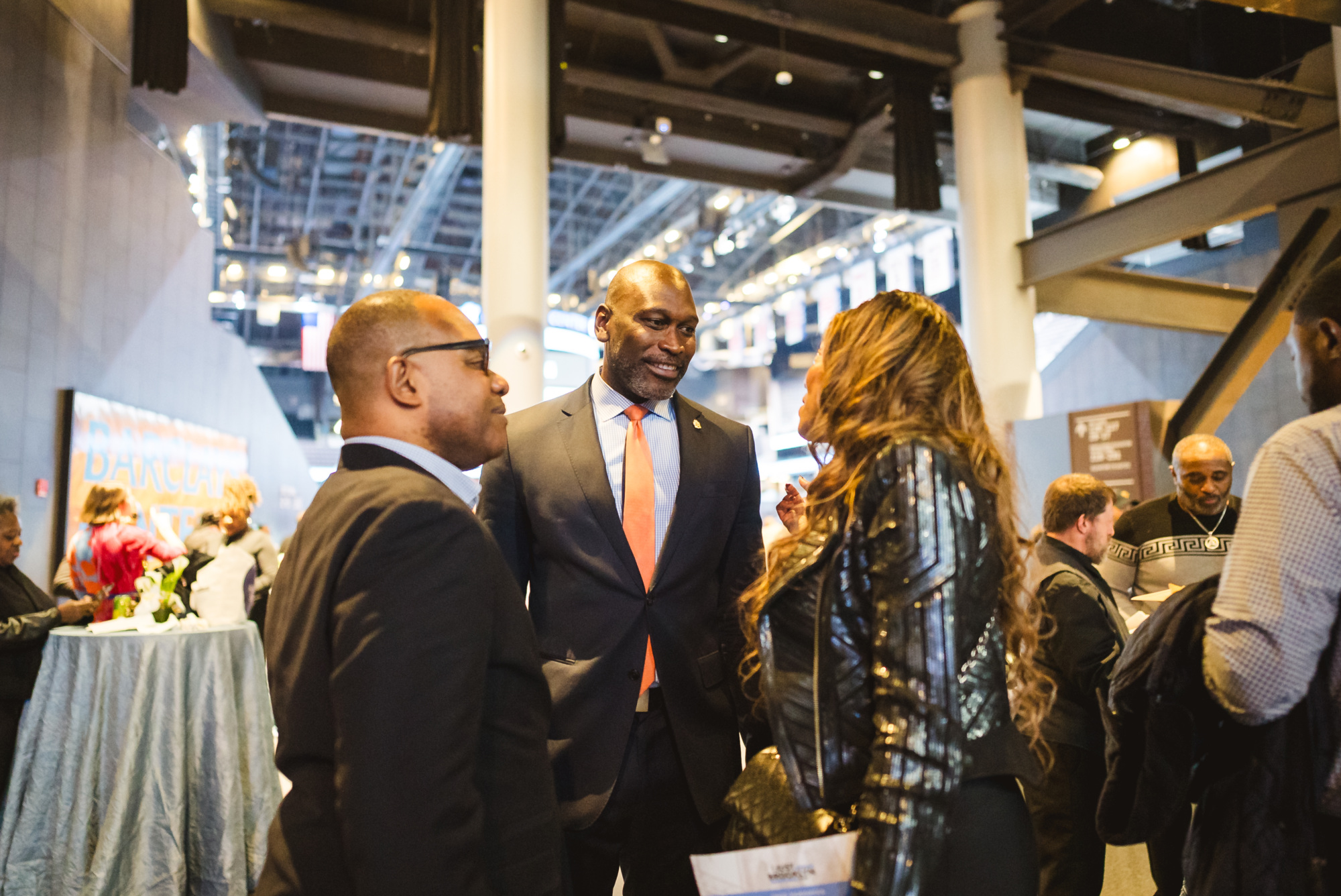 Three people in business attire converse at an indoor networking event with other attendees and tables visible in the background.