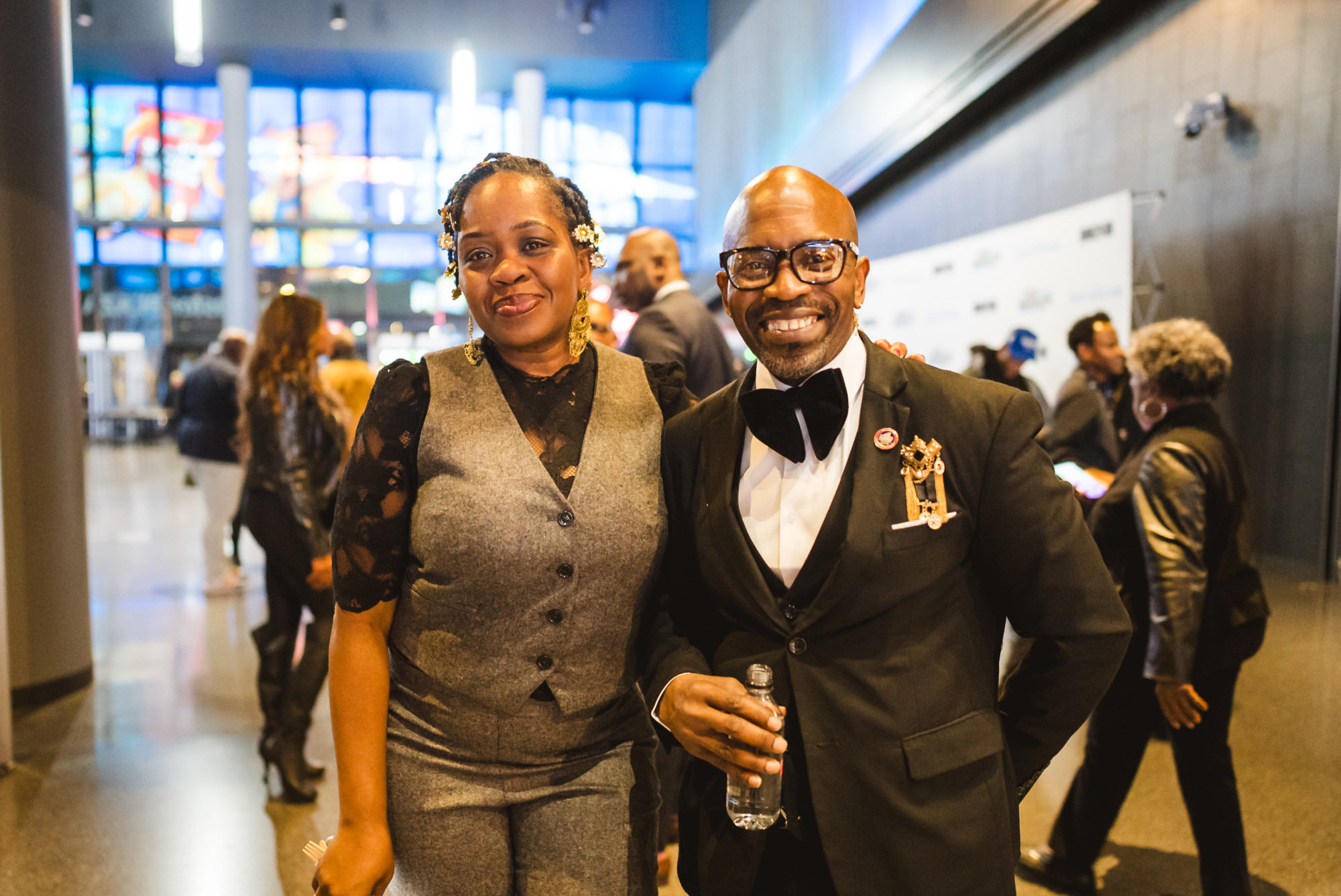 A woman and a man in formal attire pose and smile for the camera at an indoor event with other people in the background.