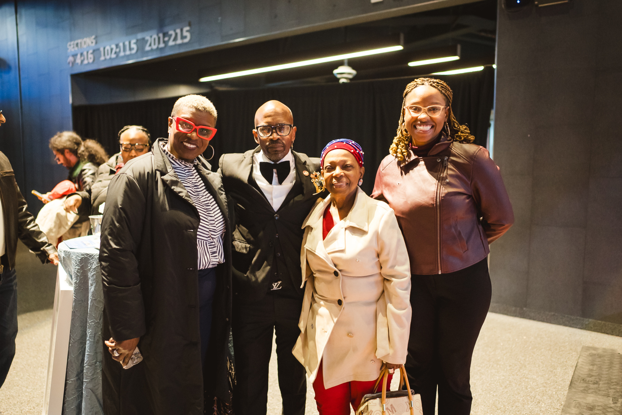Four adults pose together indoors at an event; three women and one man, all smiling and dressed in formal or semi-formal attire.