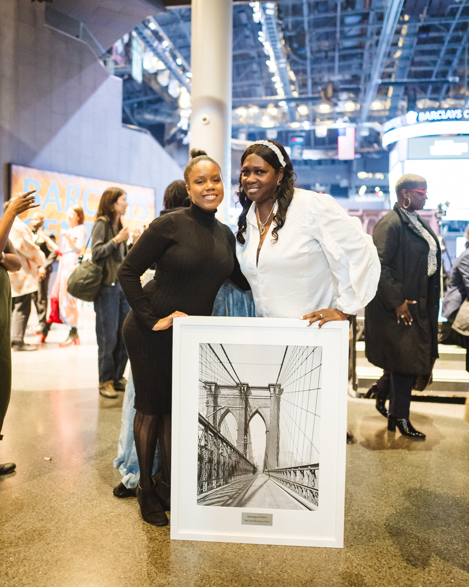 Two people pose indoors, smiling, with one holding a framed black-and-white drawing of the Brooklyn Bridge. There are other people and signs in the background.