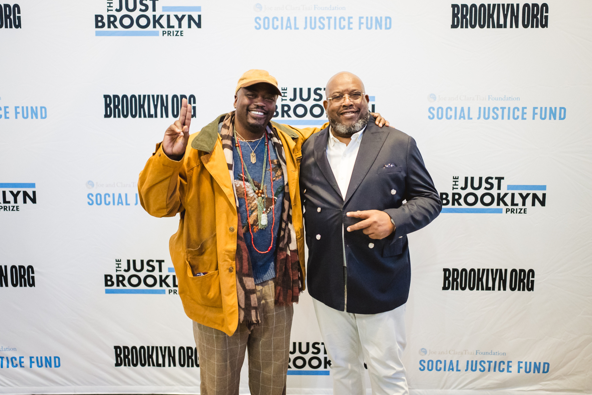 Two men stand in front of a backdrop with "Brooklyn.org," "Social Justice Fund," and "The Just Brooklyn Prize" logos. One man holds up a peace sign.