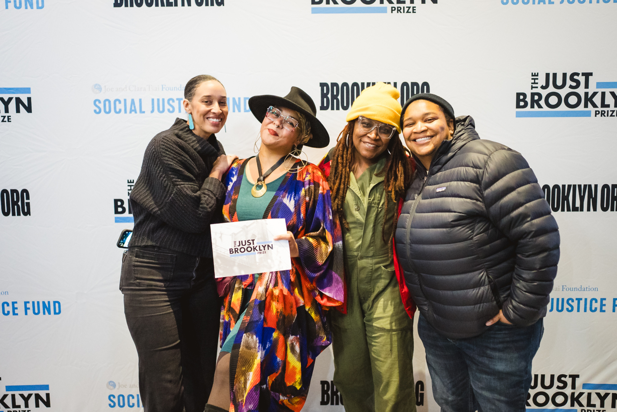 Four people stand together smiling in front of a backdrop with "Social Justice Fund" and "Just Brooklyn Prize" logos; one person holds a sign reading "Just Brooklyn.